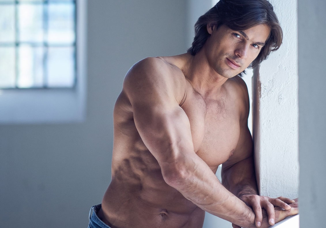 Attractive muscular shirtless guy wearing jeans posing seductively near a window.