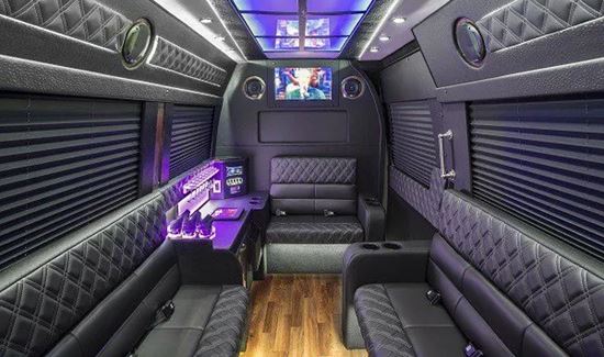 Party bus interior with black leather seating, wood flooring, purple accent lighting, and TV screens.