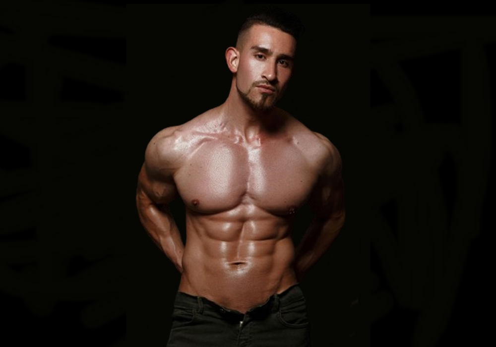 Shirtless muscular man posing against dark backdrop, with lighting highlighting defined abs, chest, and arms.