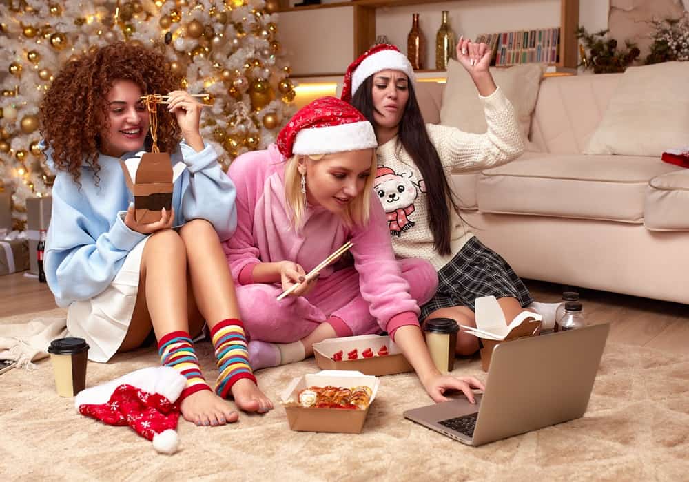 Three women enjoying a movie night seated on the floor in front of a Christmas tree wearing Santa hats and pajamas. 