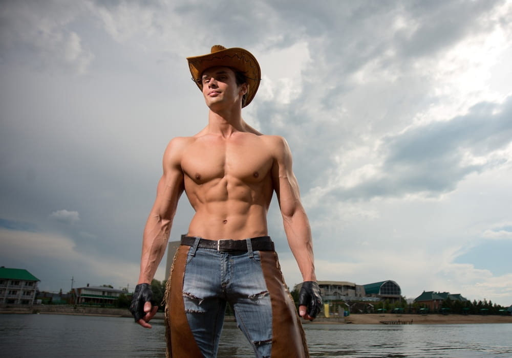 Muscular shirtless man wearing cowboy hat, gazing confidently toward the viewer.