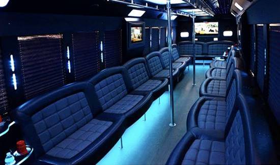 Party bus interior with dark leather seating, cool blue lighting, and central dance poles.