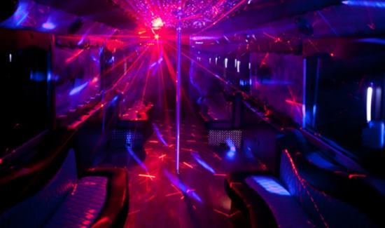 Party bus interior with vibrant red and blue laser lighting, leather seating, and a central dance pole.