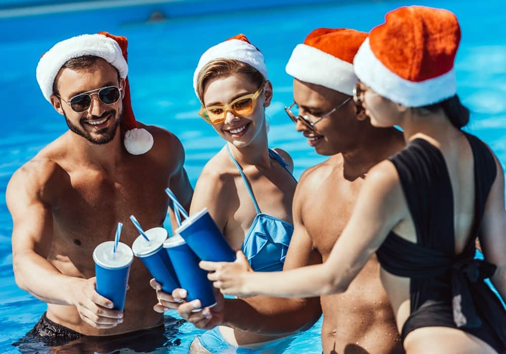 Group of young adults at a pool party wearing swimsuits and Santa hats toasting with blue cups in the pool.