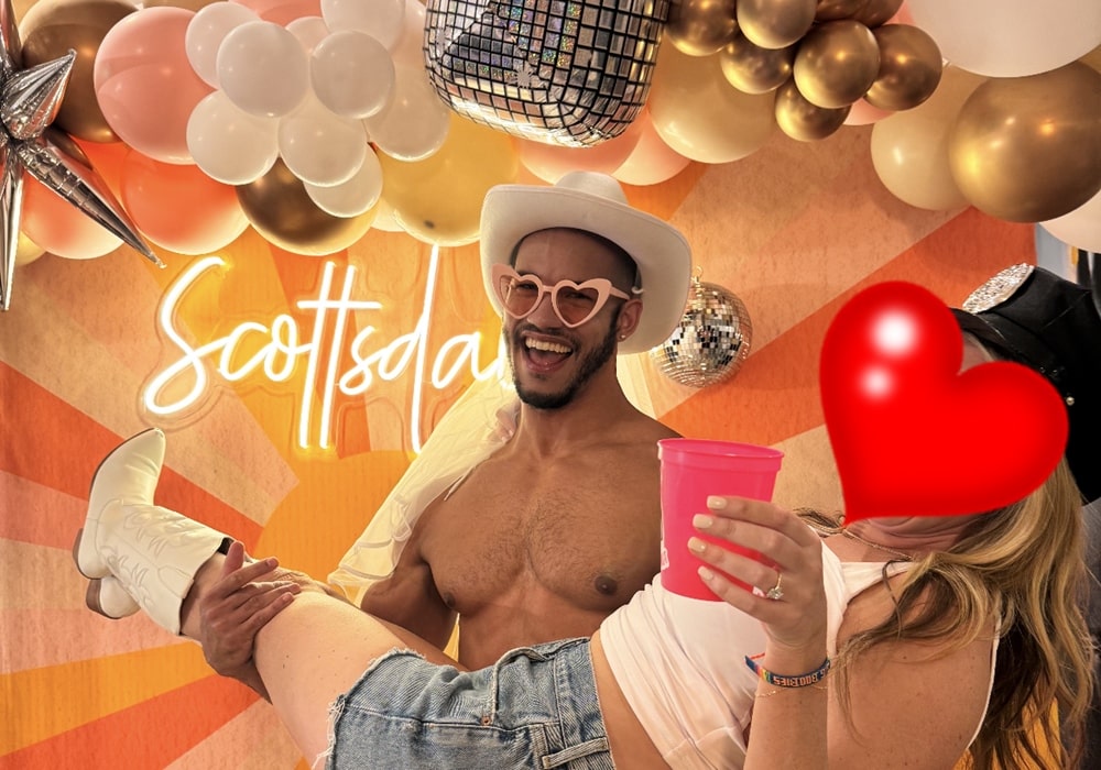 Muscular shirtless male stripper wearing sunglasses and a white cowboy hat holding a woman up against a festive background.