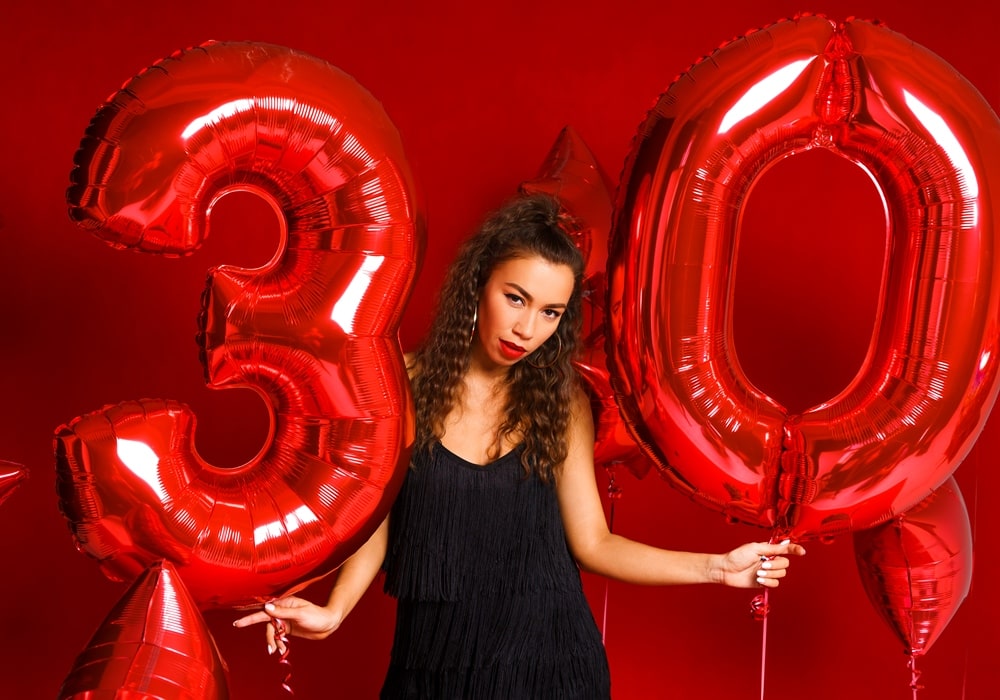 Woman wearing a black dress posing against a red background with large red balloons shaped like the numbers 3 and 0.