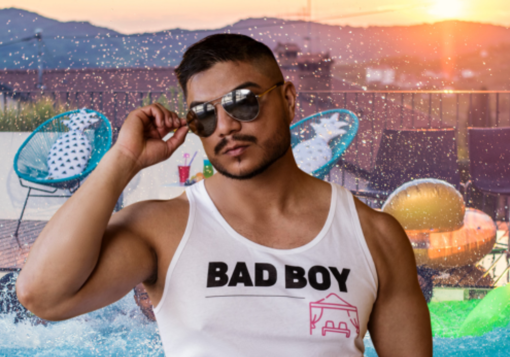 Muscular Cabana Boy wearing sunglasses and a white tank top with 'Bad Boy' text, posing in front of a poolside scene at sunset.