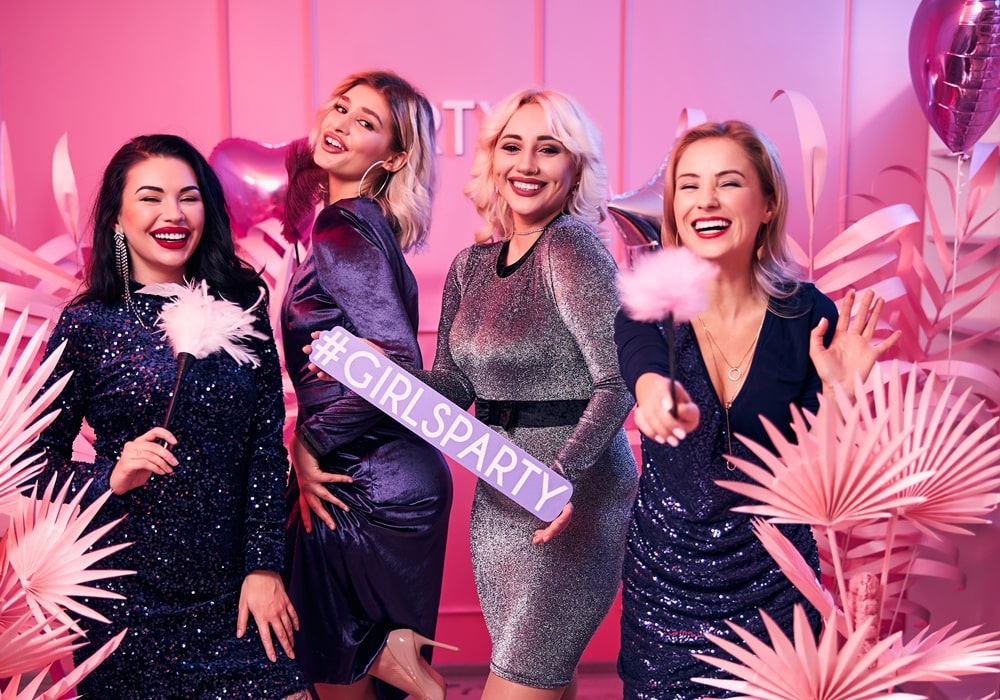 Four joyful women dressed in elegant outfits posing in a pink setting at an All Girls Party. 