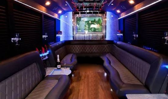 Luxury party bus interior with leather seating, wood flooring, blue lighting, and a large TV screen at the back.