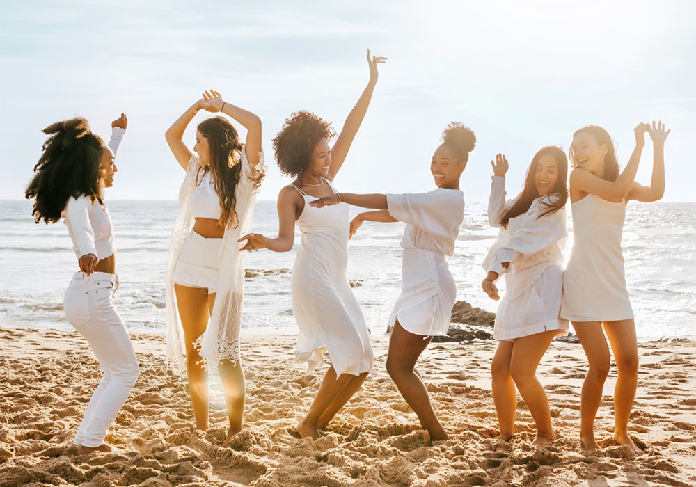Group of cheerful women all dressed in white, having fun and dancing at a beach party.