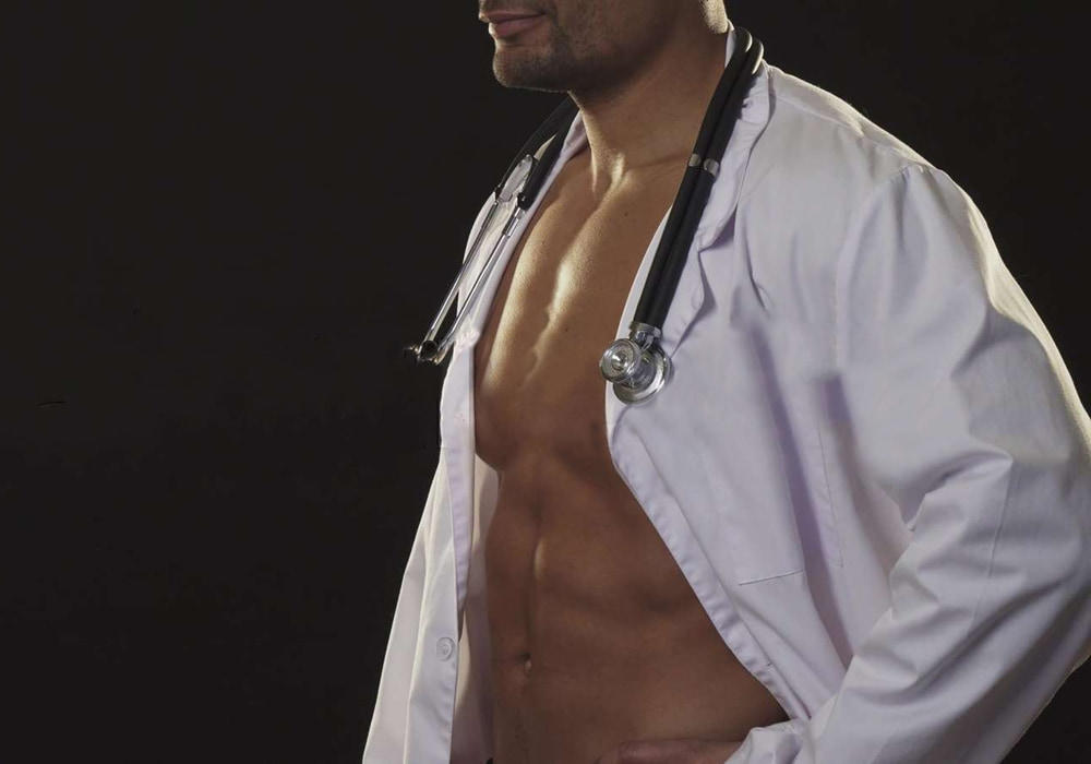 Muscular male party entertainer in white doctor coat and stethoscope, shirtless beneath, posing against a dark backdrop.