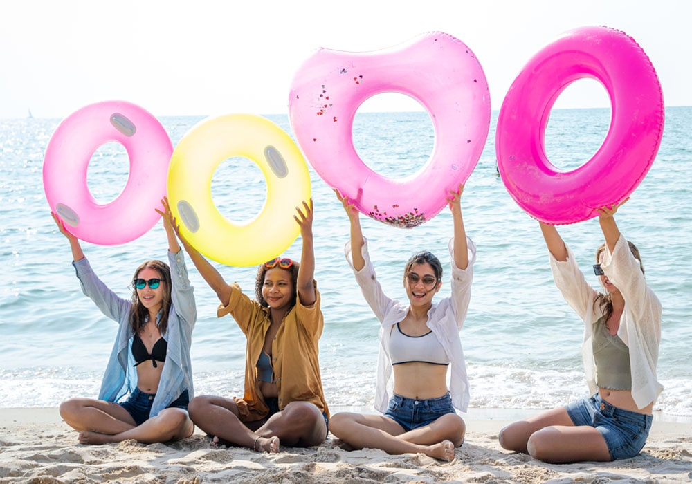 Group of young women posing at the beach holding up inflatable floats with the ocean in the background.