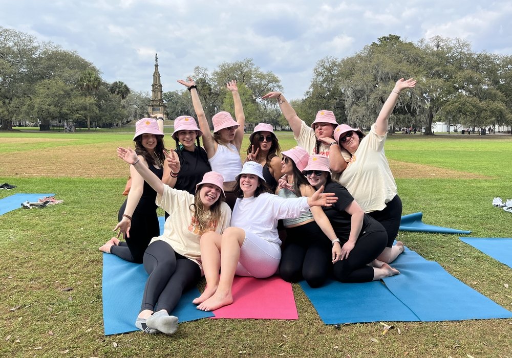 Bachelorette party group in pink bucket hats posing on yoga mats in a park, smiling with arms raised in celebration.