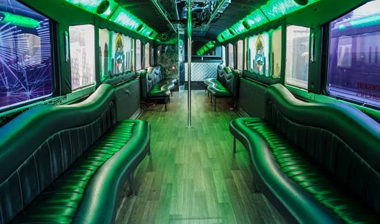 Spacious party bus interior with black leather seats on both sides, green LED lights, wood flooring, and a dance pole.