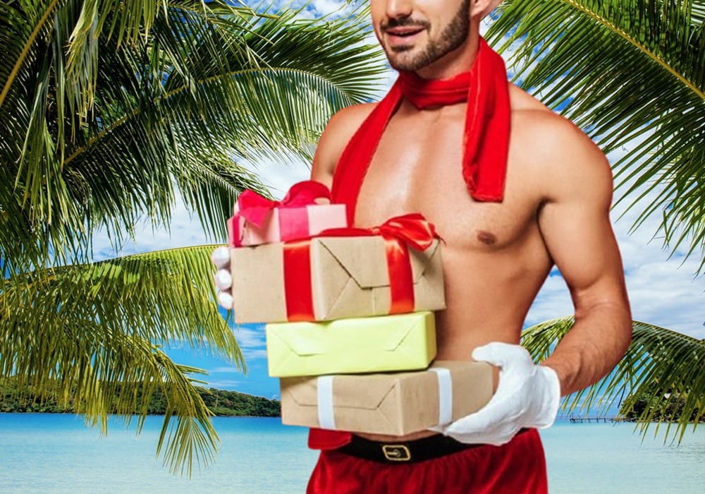 Young shirtless man dressed in Santa pants and hat holding a stack of wrapped gifts in front of a tropical setting.