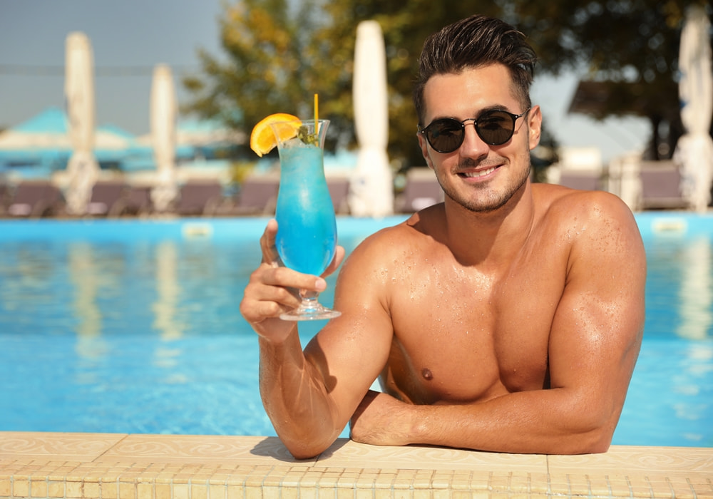 Fit, shirtless cabana boy in sunglasses smiling by the pool, holding a blue cocktail with orange garnish.