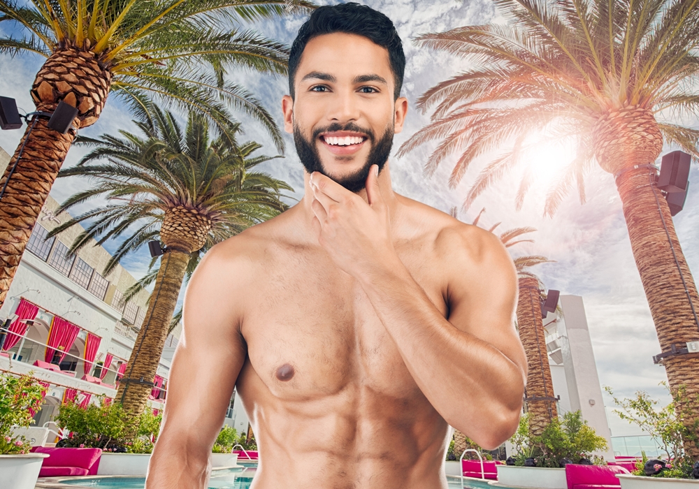 Shirtless, muscular man smiles and poses by a tropical poolside with palm trees and pink loungers.