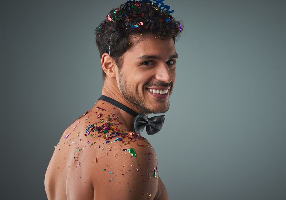  Shirtless man wearing a black bowtie smiling over his shoulder having colorful confetti in his hair, on his back.