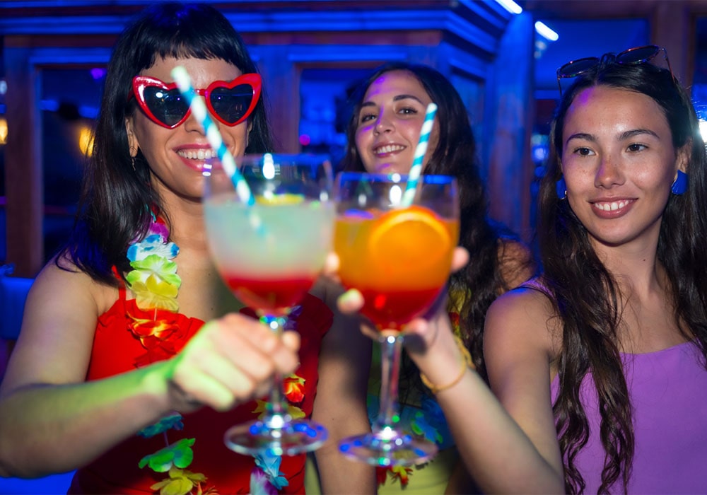 Three women enjoying a night out at a club with blue lighting smiling and toasting with colorful cocktails.