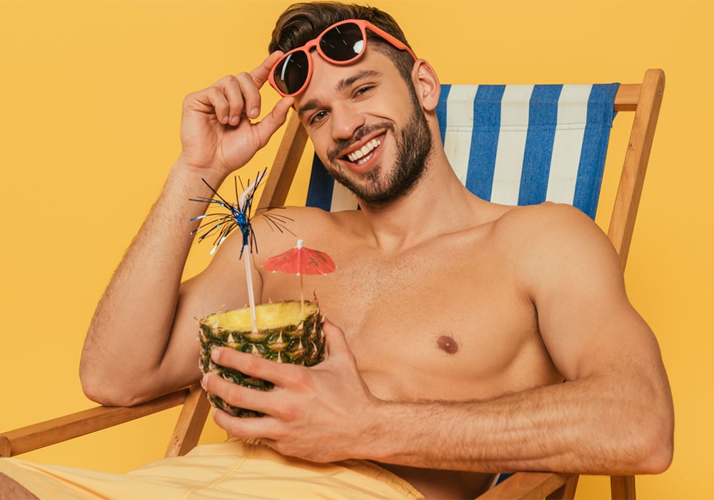 Shirtless man sitting on a striped beach chair against a yellow background holding a drink in a hollowed-out pineapple.
