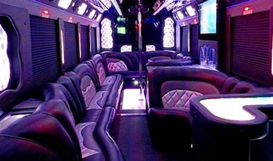 Luxury party bus interior with curved leather couches, purple ambient lighting, and a dance pole.