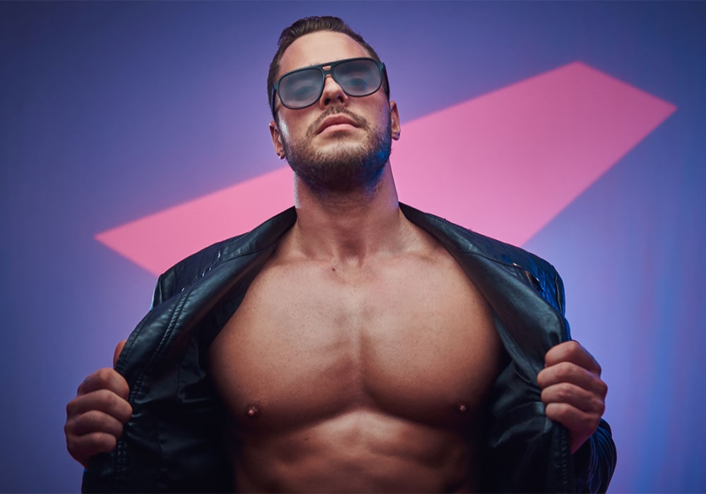Male stripper in sunglasses and leather jacket, revealing muscular chest, set against vibrant purple-blue background.