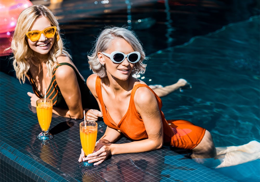 Two blondes lounging in a pool at the edge, each holding a glass of mimosa, both wearing swimsuits and sunglasses.