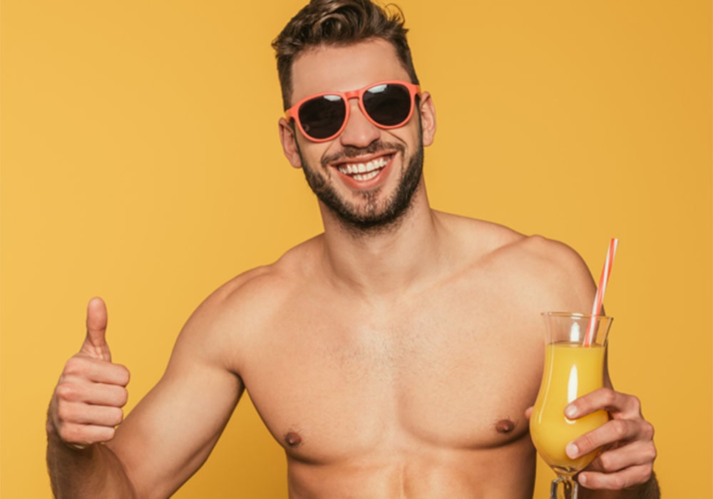Attractive shirtless guy smiling and posing in front of a yellow background holding a cocktail.