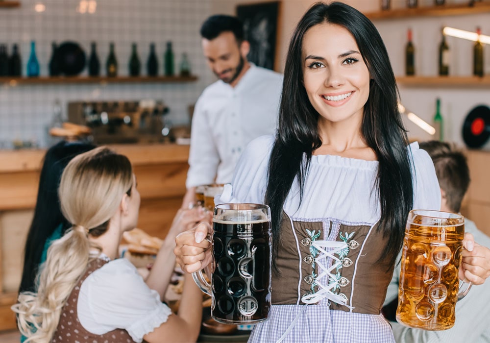 Brunette in a Bavarian-style outfit holding two beer mugs with people in the background at an Oktoberfest party.