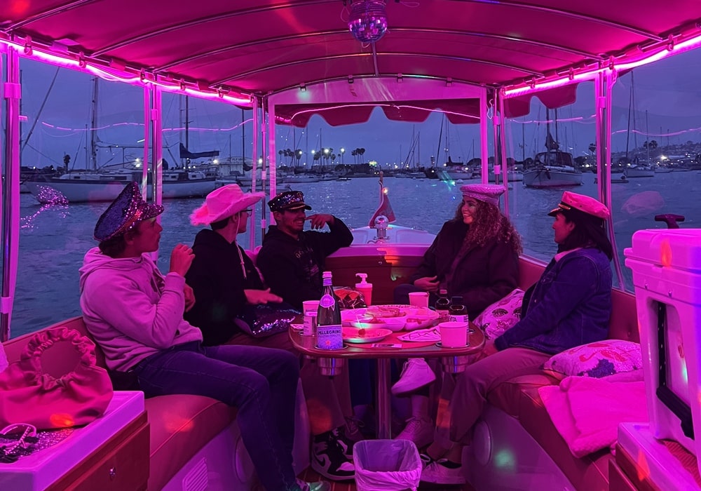 Five friends celebrate a bachelorette party on a pink-lit boat with drinks, snacks, party hats, and a disco ball.