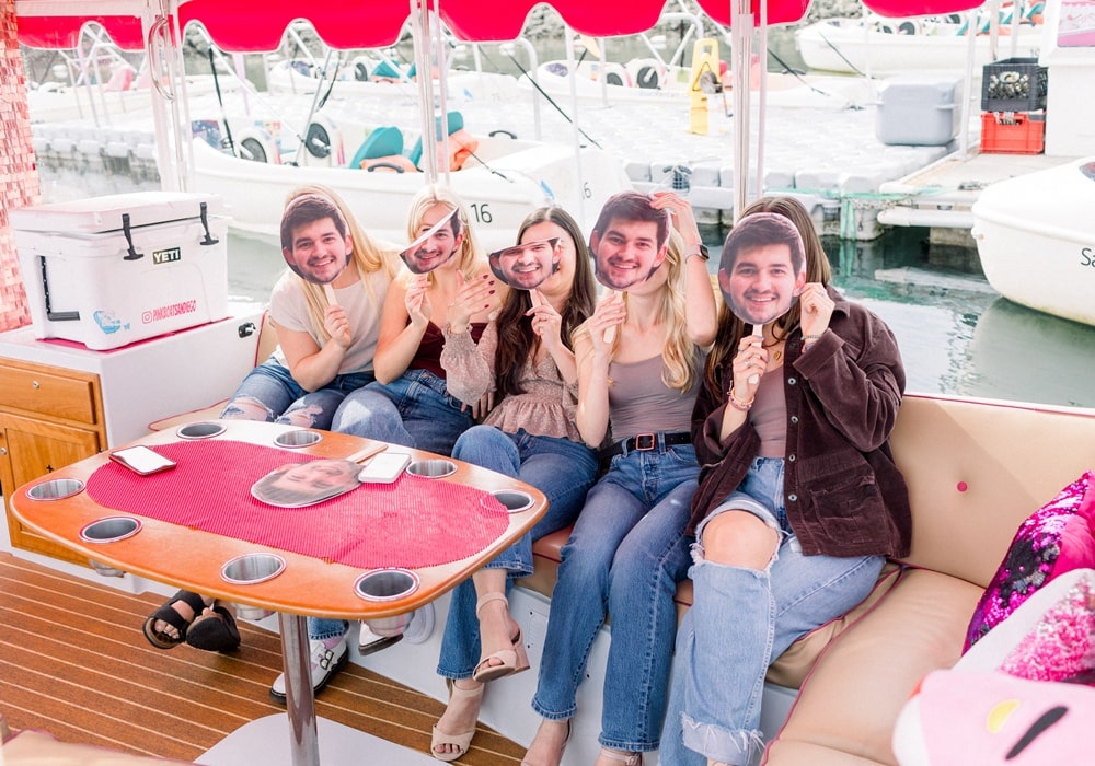 Group of women at a bachelorette party on a San Diego pink boat, holding masks with the groom’s face.