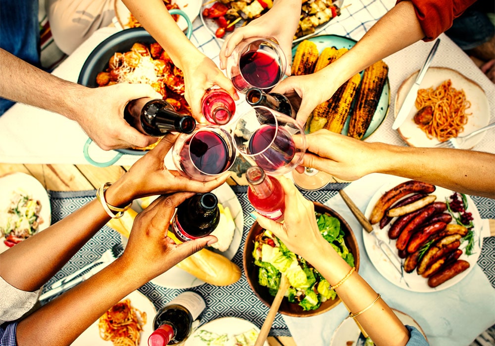 People toasting with glasses of red wine and bottles, gathered around a table filled with foods.