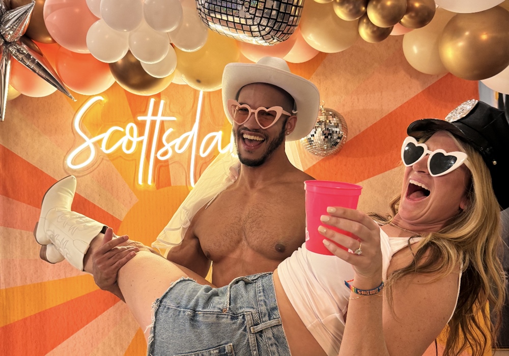 Bride-to-be is held by a cowboy-costumed stripper at a bachelorette party with a vibrant disco-western backdrop.