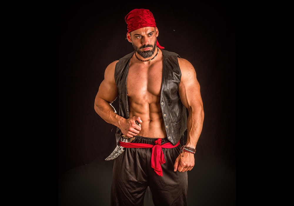 Muscular man in a pirate-themed costume with red bandana, black pants, and vest, posing while holding a dagger.