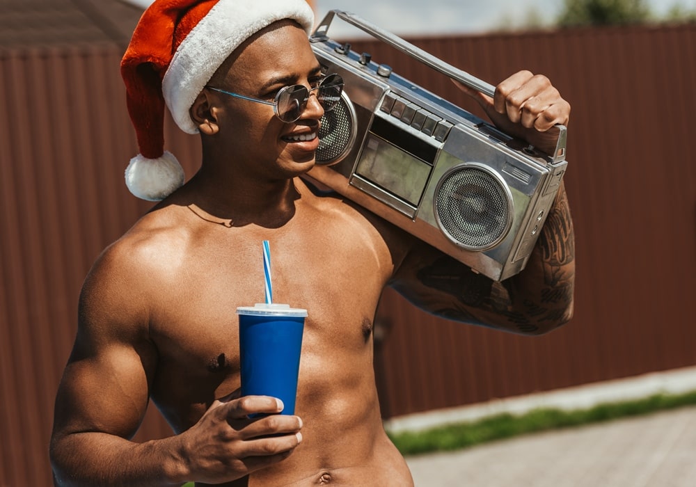 Sexy Muscular shirtless man outdoors wearing a Santa hat and sunglasses holding a large vintage boombox and a blue cup.