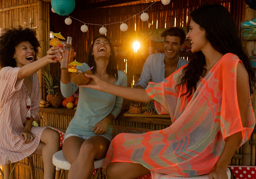 Three women laughing and drinking tropical cocktails at a tiki bar being served by a handsome male bartender. 
